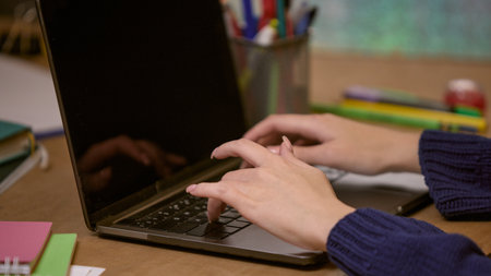 Close-up of hands typing on a laptop at a desk with office supplies and notebooks, representing a...