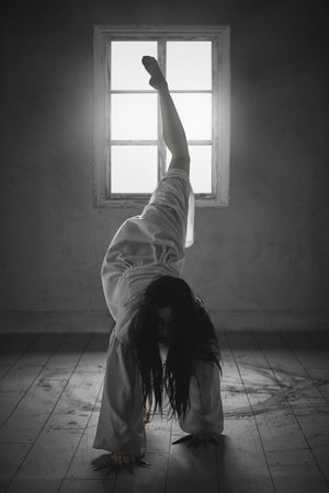 Woman dressed in white gown with flowing black hair, crouching in an eerie, animalistic pose on wooden floor with a single window behindの写真素材
