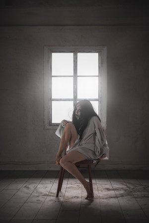 A woman with black hair, white skin and dark eyes sits on chair win white gown in a terrifying posture, evoking the sense of horror and mysticismの写真素材