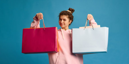 Beautiful, happy young girl, posing raising hands holding shopping bags with copy space against blue...
