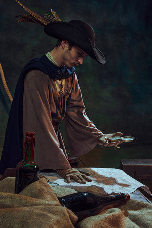 Focused man pirate captain in feathered hat and gloves, looking at vintage map and compass, against...