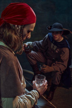 Focus on man, pirate in red bandana standing with thoughtful face with glass of rum, with his mate...