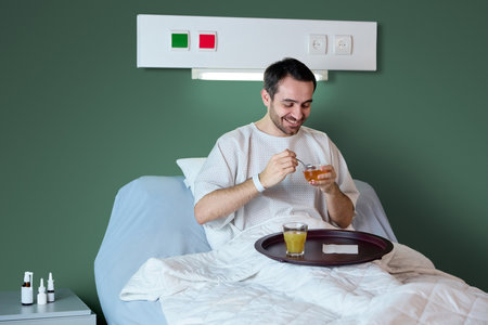 Male patient in white hospital gown sitting in bed with food tray, smiling, eating and drinking juice against green wall background.の写真素材