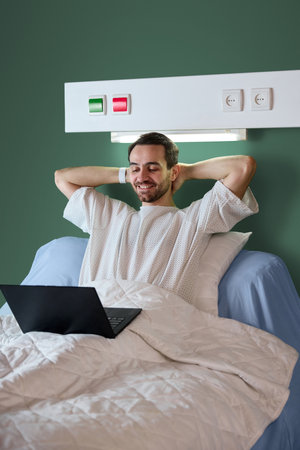 Happy smiling man, patient sitting in bed with blanket, in hospital gown and joyfully watching movie on laptop against green wall background.の写真素材