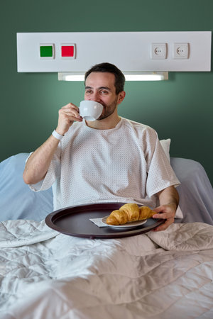 Joyful dark-haired adult man, patient in hospital gown sitting in bed, drinking coffee with croissant of food tray against green wall. Positive recoveryの写真素材