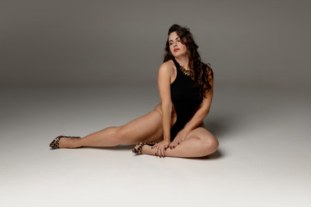 Confident woman, model, posing elegantly sitting on floor and demonstrating beauty of her body curves against grey studio background.の写真素材