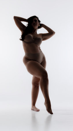 Silhouette of woman standing gracefully in lingerie, arms raised behind head, exuding elegance and empowerment against white studio background.の写真素材