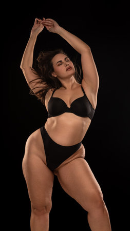 Overweight young woman in black underwear, posing in relaxed posture with closed eyes and holding arms raised against black studio background.の写真素材
