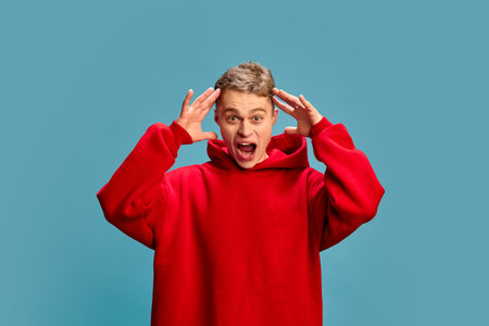 Portrait of young attractive man with surprised expression, open mouth and putting hands on head...