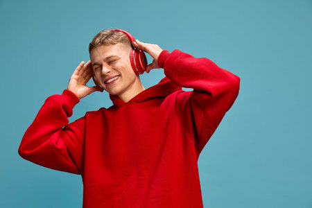 Young handsome man, dressed casual, listening music in headphones, expressing relax and joy against blue studio background.の写真素材