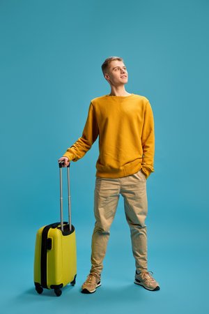Full length photo of young man, dressed casually stands ready to adventures with luggage against blue studio background.の写真素材