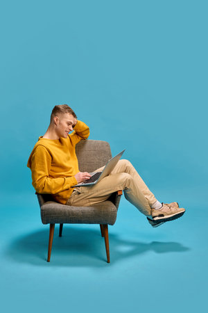 Man sitting on chair working on laptop, leaning back with relaxed posture, focusing intently on screen, conveying engagement and productivity against blue background.の写真素材