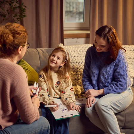 Young girl shares joyful moment with psychologist while drawing, her smiling mother observes drawing warmly, creating supportive therapy setting.の写真素材