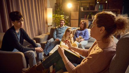 Psychologist takes notes, listening to teen boy expressing his thoughts and feelings, family members sitting in background.の写真素材