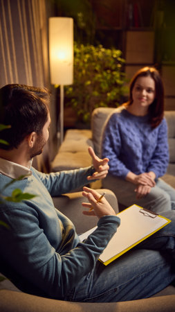 Psychologist gesturing actively while speaking to a young woman, both seated in a cozy room with warm lighting and plants in the background.の写真素材