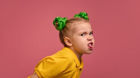 Child, girl with cheeky smirk, sticking out tongue playfully, wearing yellow shirt and green braided buns against pink studio background.の写真素材