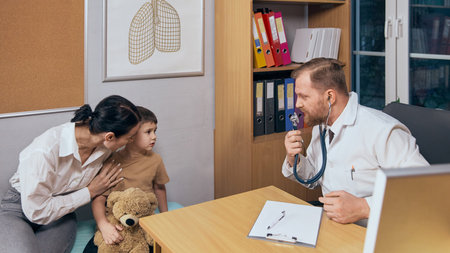 Male pediatrician with beard using stethoscope while examining preschool boy against organized office background. Child came to doctor with his motherの写真素材