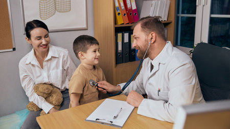 Smiling boy with mother visiting doctor, pediatrician listens to boy heartbeat using stethoscope....