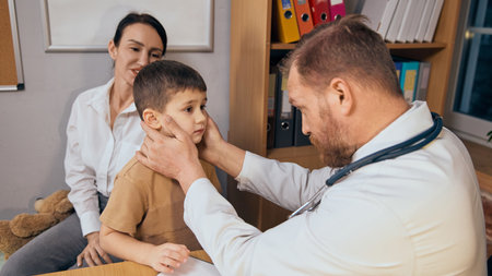 Male doctor inspecting young boy neck, lymph nodes with both hands during medical consultation. Attentive mother sitting in backgroundの写真素材