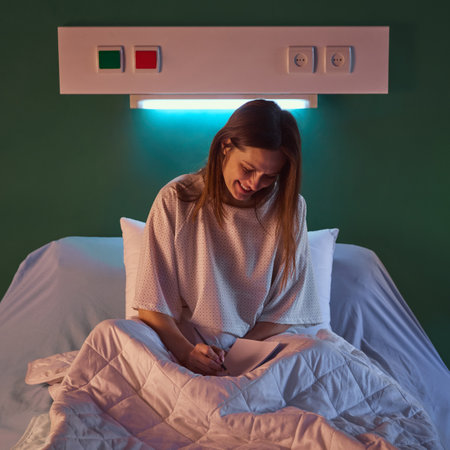 Young woman sitting in bed in her hospital ward in evening and making notes in notebook against...