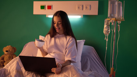Smiling teen girl sitting on bed in hospital gown with her laptop, drinking tea and watching...