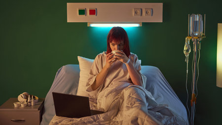 Young red-haired woman sitting on bed, sipping tea and watching movie on her laptop in hospital ward...