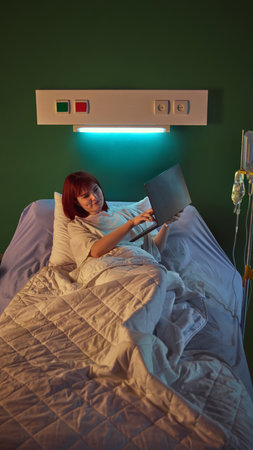 Young smiling red-haired woman lying in hospital bed, holding laptop and having online video call...