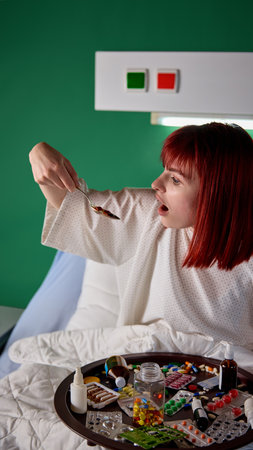 Young red-haired woman sitting in bed with tray filled with many different meds and taking pills with large spoon. Recovery process in modern clinicの写真素材
