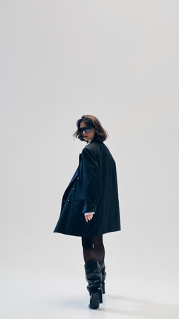 Full-length of confident businesswoman in oversized black blazer, leather boots and sunglasses, posing in motion against light studio backgroundの写真素材