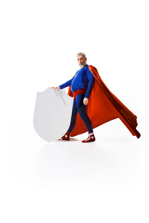 Full-length portrait of grey-haired bearded man, with belly, in superhero costume with red cape standing with white shield against white studio backgroundの写真素材