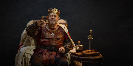 Victory. Senior man, medieval king in crown sitting on throne and raisin glass of wine against black studio backgroundの写真素材