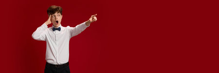 Young emotional boy in white shirt and bow tie emotionally pointing with hand, expressing shock and excitement against red studio backgroundの写真素材