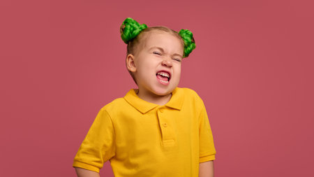 Portrait of emotive preschool girl, child in yellow shirt and braided buns, making funny faces, grimacing, playing against pink studio background.の写真素材