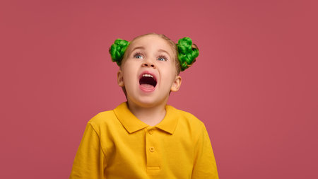 Portrait of adorable child, preschool girl wearing yellow polo shirt, with braided buns hairstyle emotionally grimacing against pink studio background.の写真素材
