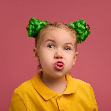 Portrait of playful child, preschool girl in yellow shirt and braided buns hairstyle emotionally making funny faces against pink studio background.の写真素材