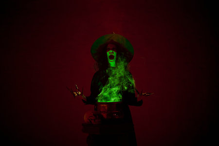 Shouting witch in black cloak and pointed hat making spell with bubbling cauldron and green mist rising around, in a dimly lit chamber.の写真素材