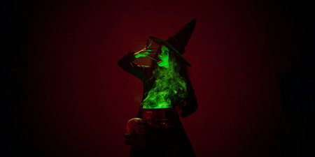 Portrait of woman, witch in black cloak and pointed hat, standing next to bubbling cauldron with green mist against dark red background.の写真素材