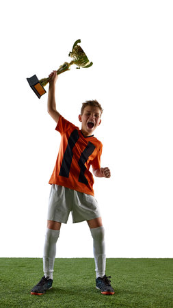 Full-length image of boy, soccer player in orange jersey and white shorts, standing on grassy surface, lifting trophy. Victoryの写真素材