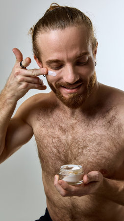 Shirtless man with beard smiling while applying white face cream, embracing self-care with cheerful expression against white studio background.の写真素材