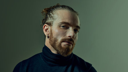 Portrait of handsome man with tied up hair and beard, wearing black turtleneck, posing with...