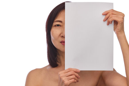 Portrait of beautiful Korean mature woman with well-kept healthy skin covering half of her face with blank paper isolated on white backgroundの写真素材