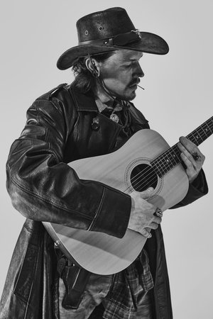 Black and white portrait of stylish man in leather coat and cowboy hat, playing guitar. Country...