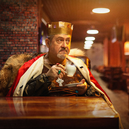 Medieval king in golden crown, fur-lined cloak, and regal robe, sitting at wooden table in warmly...