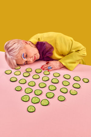 Elderly woman in yellow blazer resting head on table covered with cucumber slices, appearing peaceful against yellow background.の写真素材