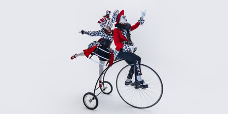 Circus duo riding vintage bicycle in synchronized movement, dressed in eccentric black, white, and red costumes over white background.の写真素材