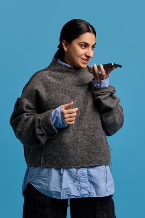 Cheerful young woman with dark hair, wearing gray sweater, holding smartphone, speaking voice...