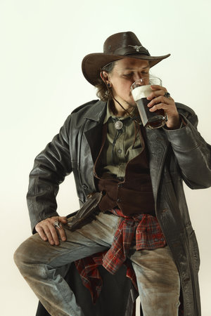 Portrait of man, cowboy in leather coat, hat, jeans and shirt drinking dark foamy beer against light studio background.の写真素材