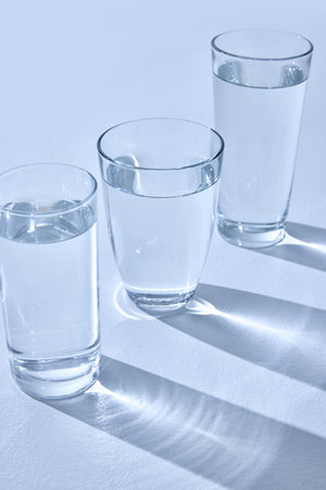 Three glasses filled with water stand on light surface, casting long, blurry shadows. Light refracts through water, creating sparkling effect. Mock up.の写真素材