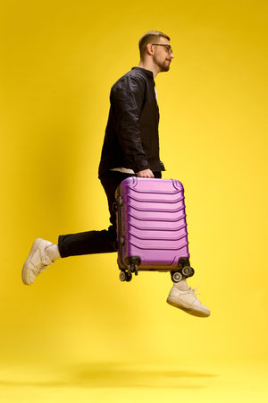 Dynamic man jumping mid-air with suitcase, dressed in black, playful motion on yellow studio background.の写真素材