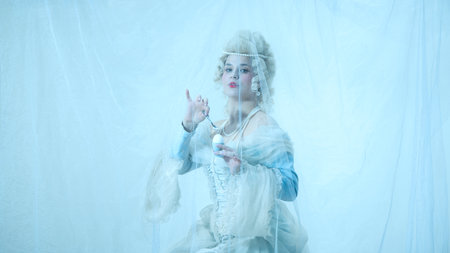 Elegant young woman in royal costume delicately cracking egg with spoon against light blue background.の写真素材